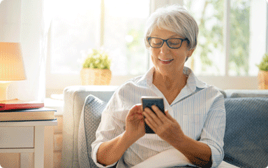 Mujer con gafas y pelo canoso mirando su smartphone sentada en el sofá.